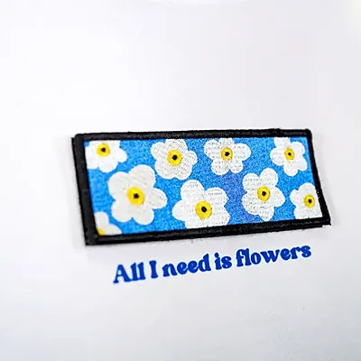 Écusson brodé rectangulaire à motif de fleurs blanches sur fond bleu, cousu sur un tee-shirt blanc